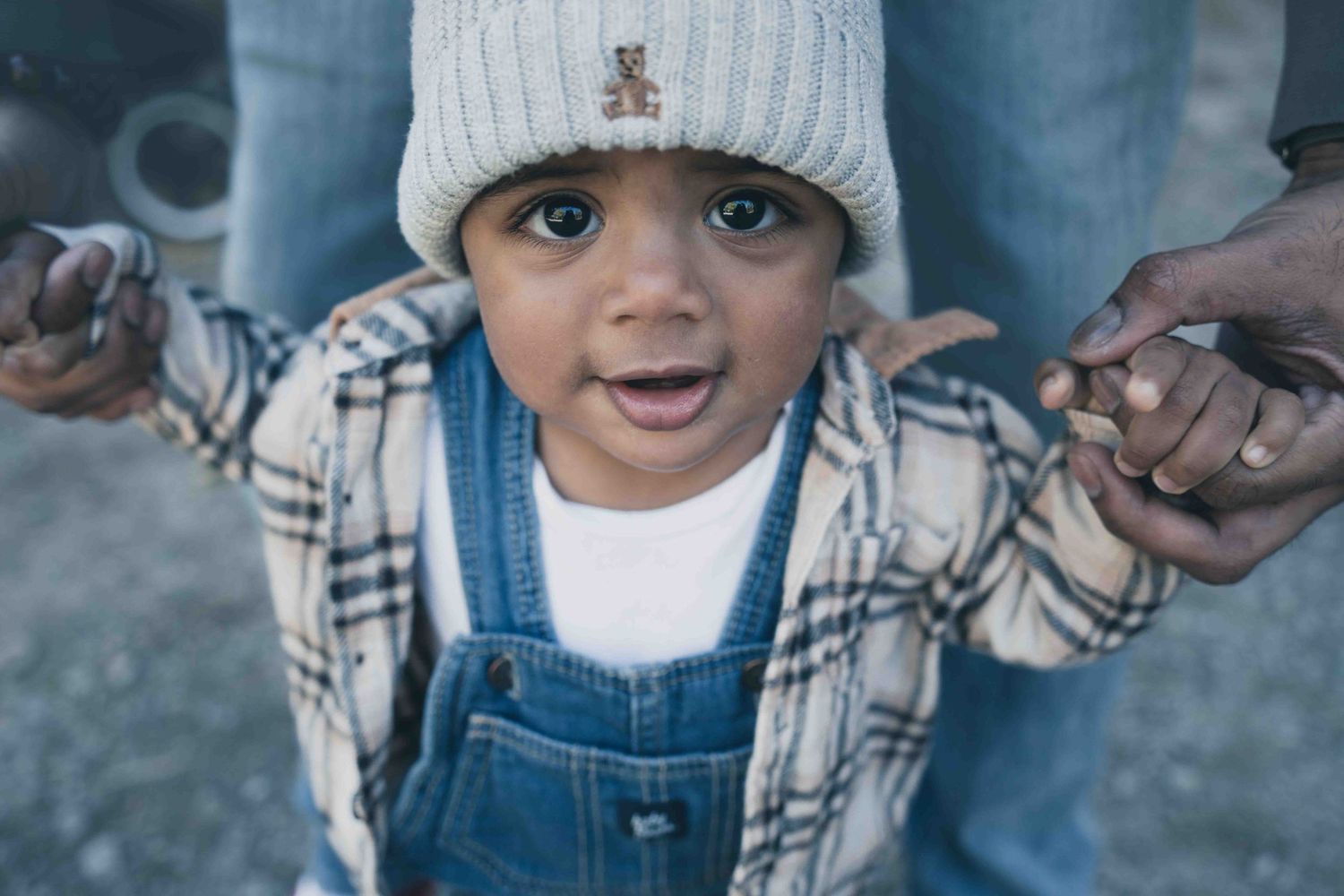 A toddler in denim overalls and a white knit hat smiles while holding an adult's hand outdoors.