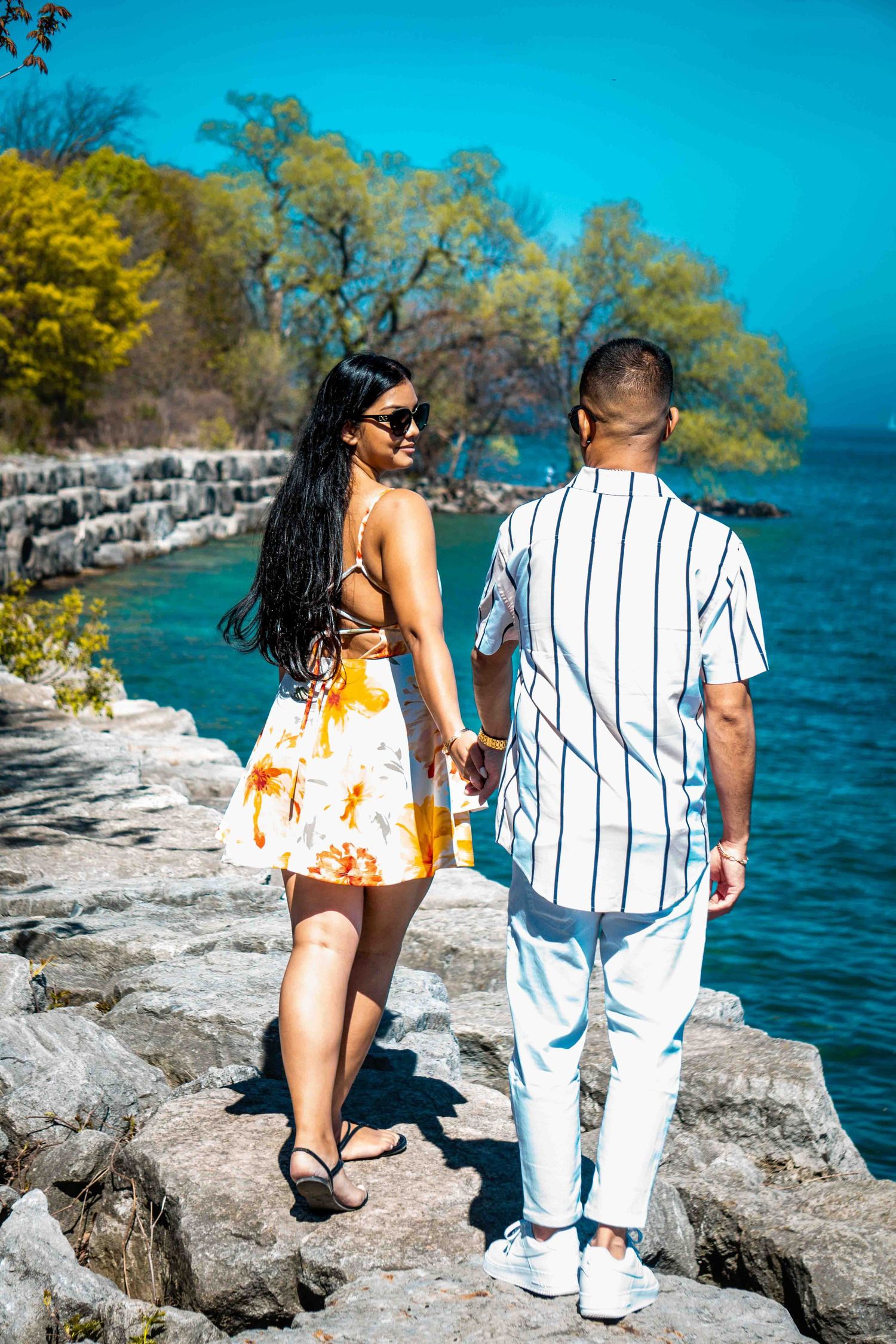 Couple walking along rocky shoreline with bright blue water in background.
