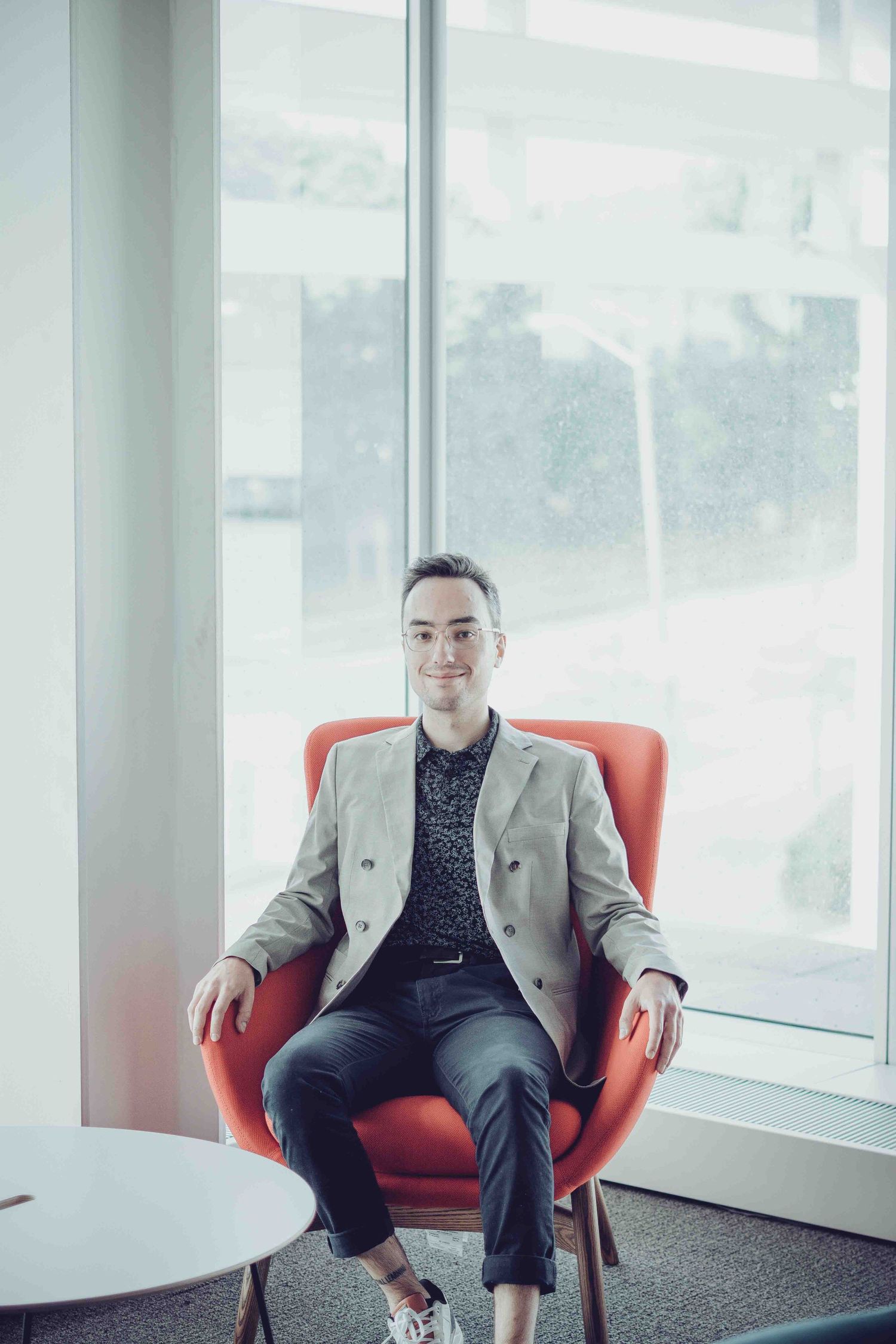 Professional in light blazer seated in red armchair against bright window backdrop.
