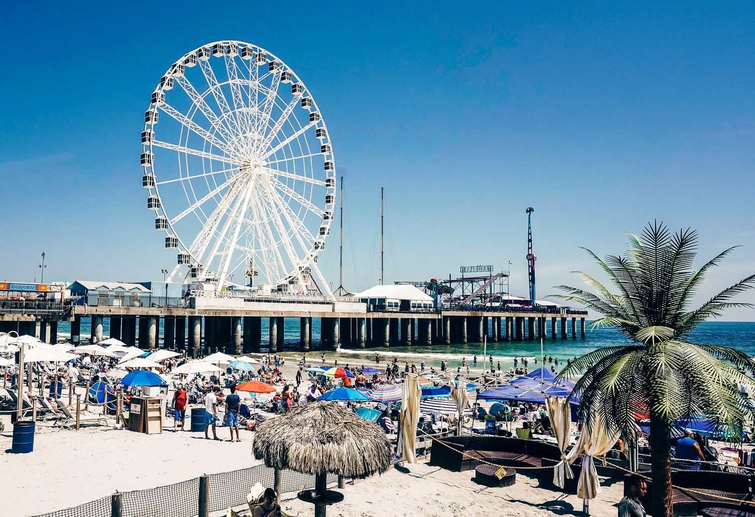Panoramic beach boardwalk scene with Ferris wheel and pier attractions on a sunny day.