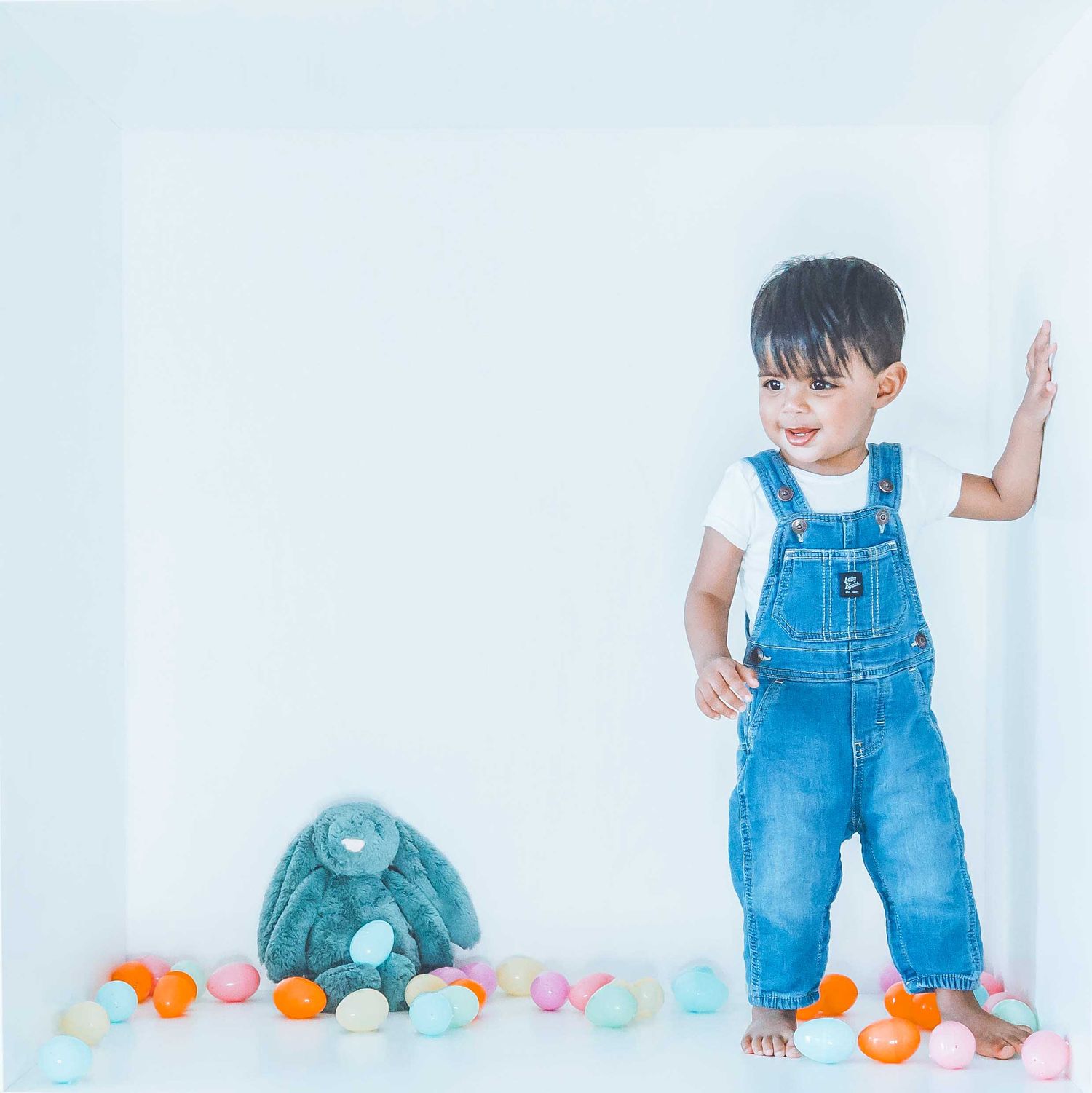 Toddler in blue overalls playing with colorful Easter eggs on white background.