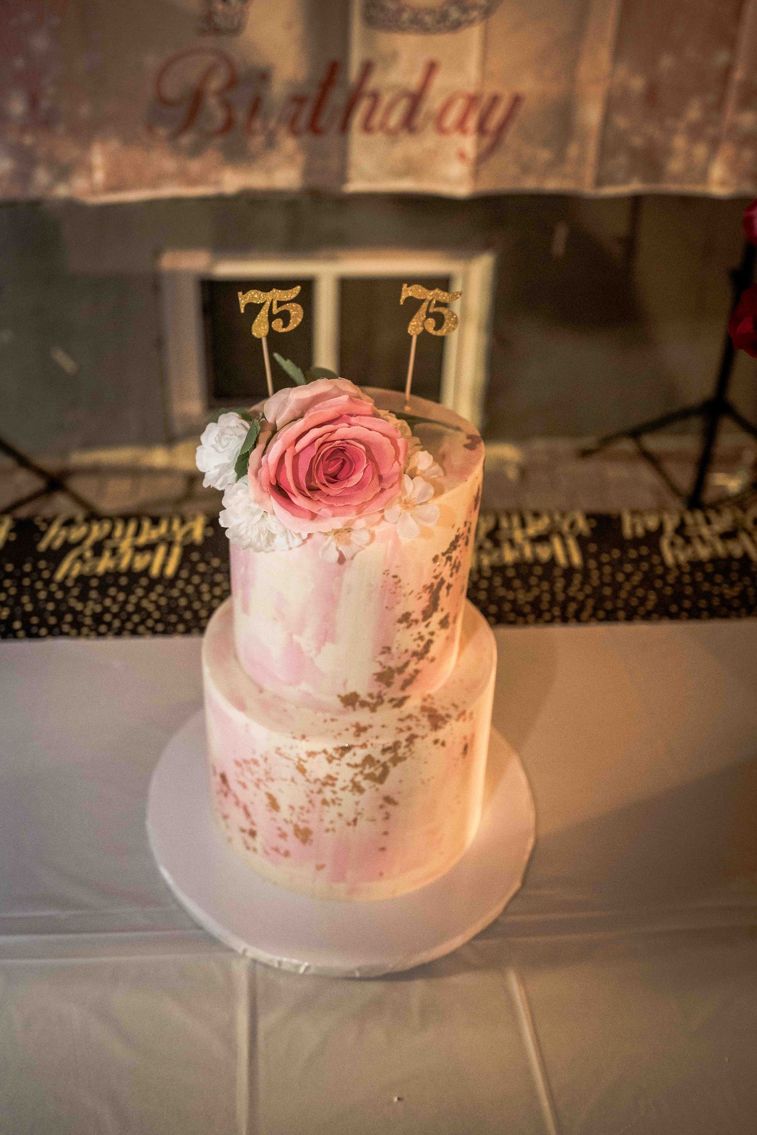 White and pink birthday cake with gold number 75 toppers on a display stand.