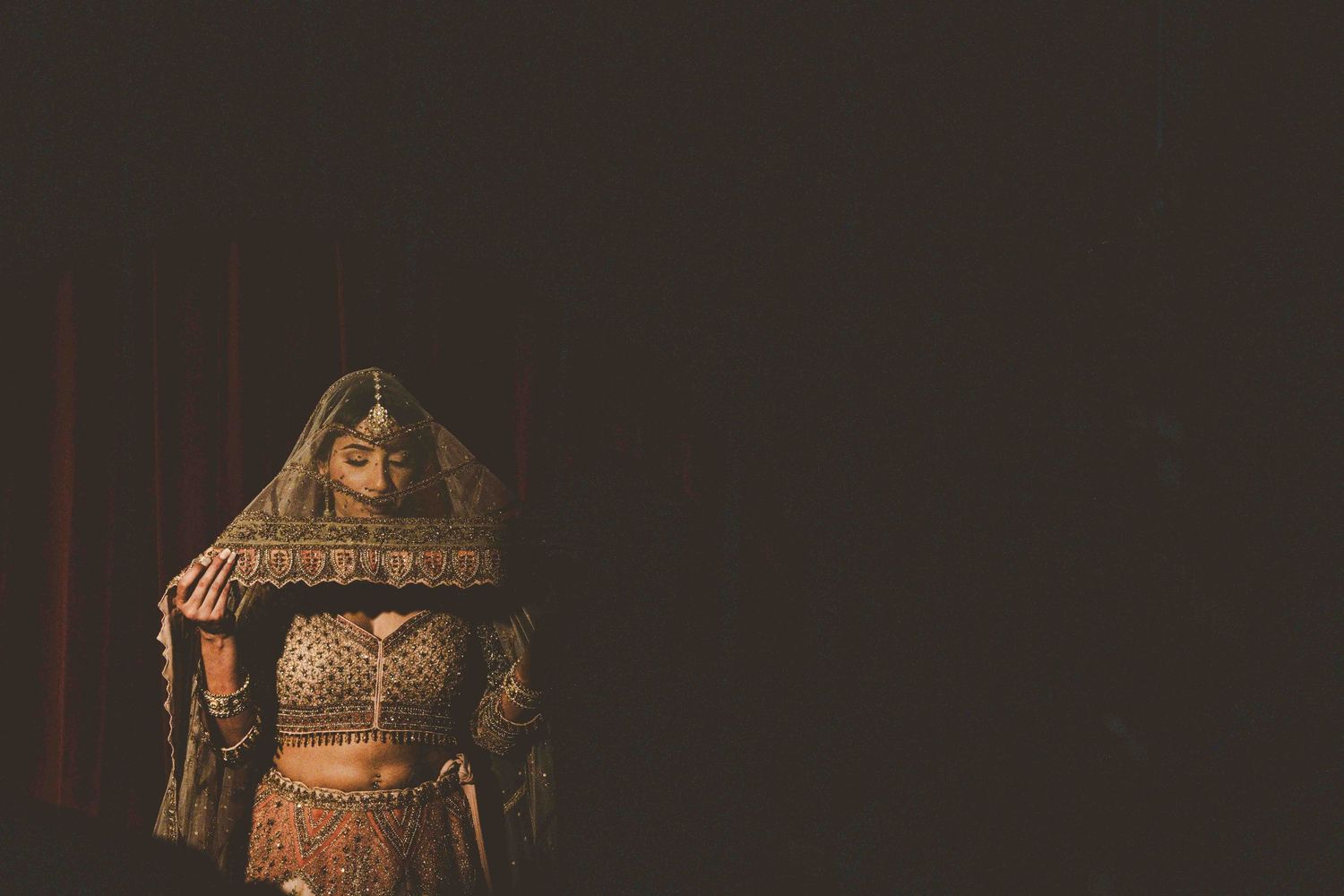 Traditional Indian bride in red and gold lehenga with ornate jewelry stands in dramatic lighting.