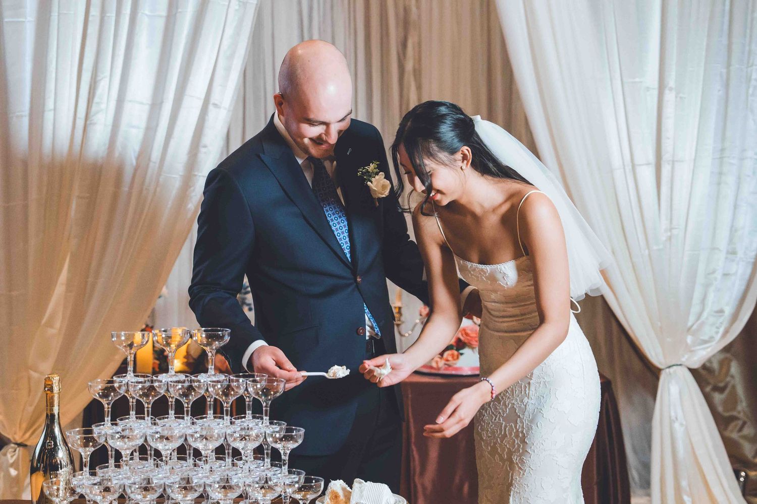 A couple stands close together near a tower of champagne glasses at an elegant indoor event.