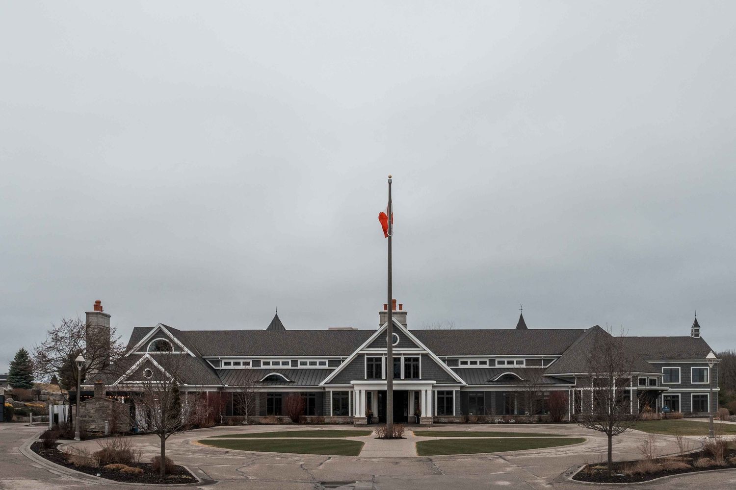 Large suburban clubhouse with red flag on tall flagpole under overcast sky, featuring gray roof and circular driveway.