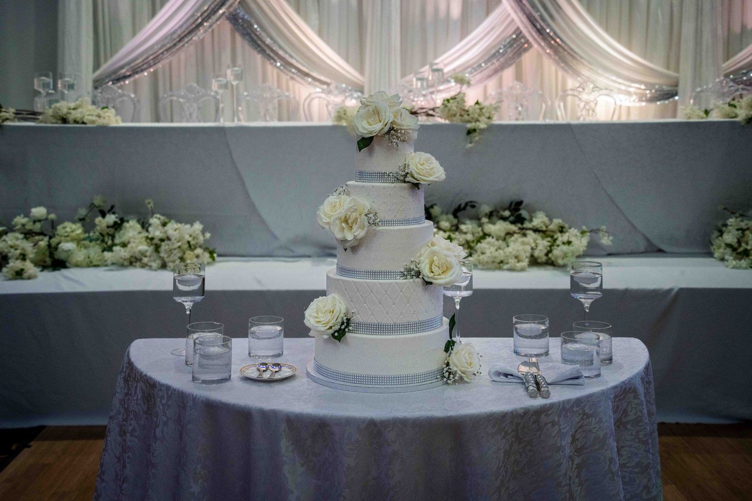 Elegant four-tier white wedding cake decorated with roses on a silver tablecloth at a reception venue.