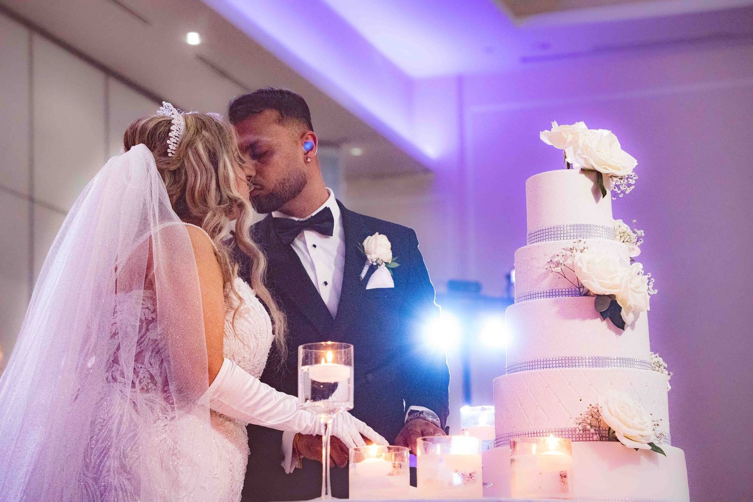 Newlyweds cutting their elegant white wedding cake under purple lighting.