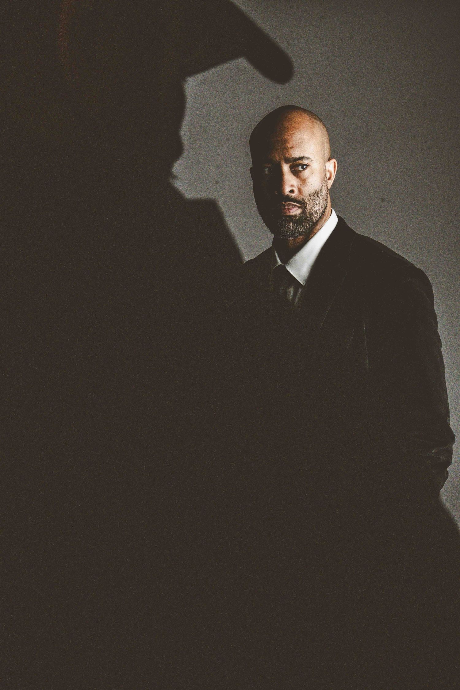 A dramatic portrait in dark lighting shows a bald man with a beard wearing a black suit and tie against a shadowy wall.