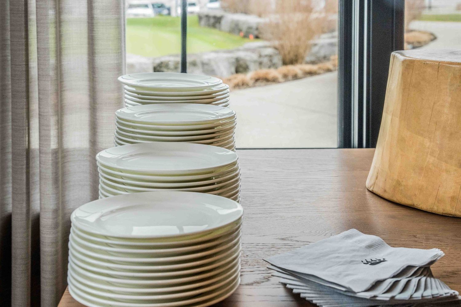 Stacks of white ceramic dinner plates on a wooden table beside folded napkins near a large window.