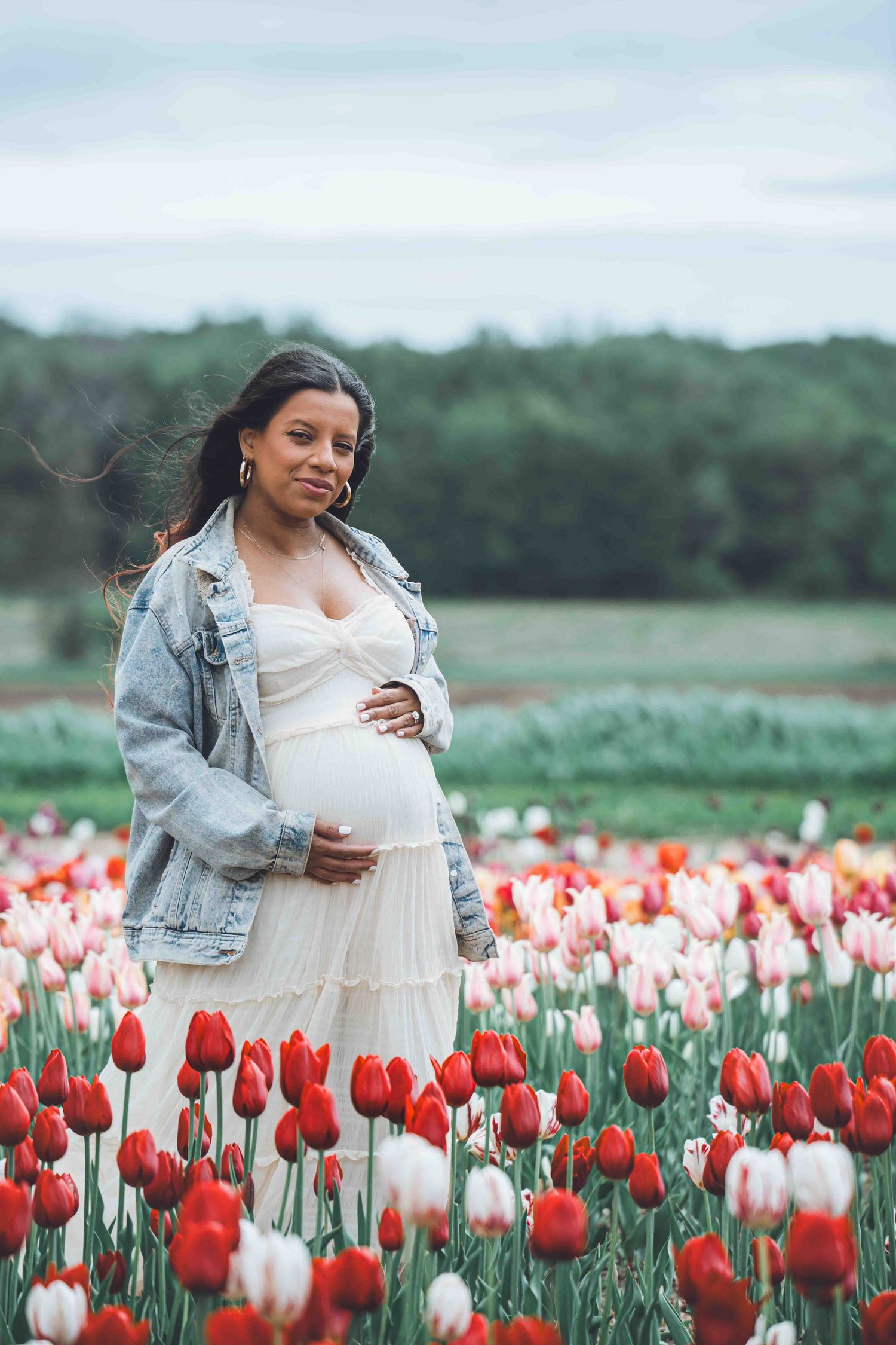 Expectant mother in white dress and denim jacket poses in vibrant tulip field during spring photoshoot.