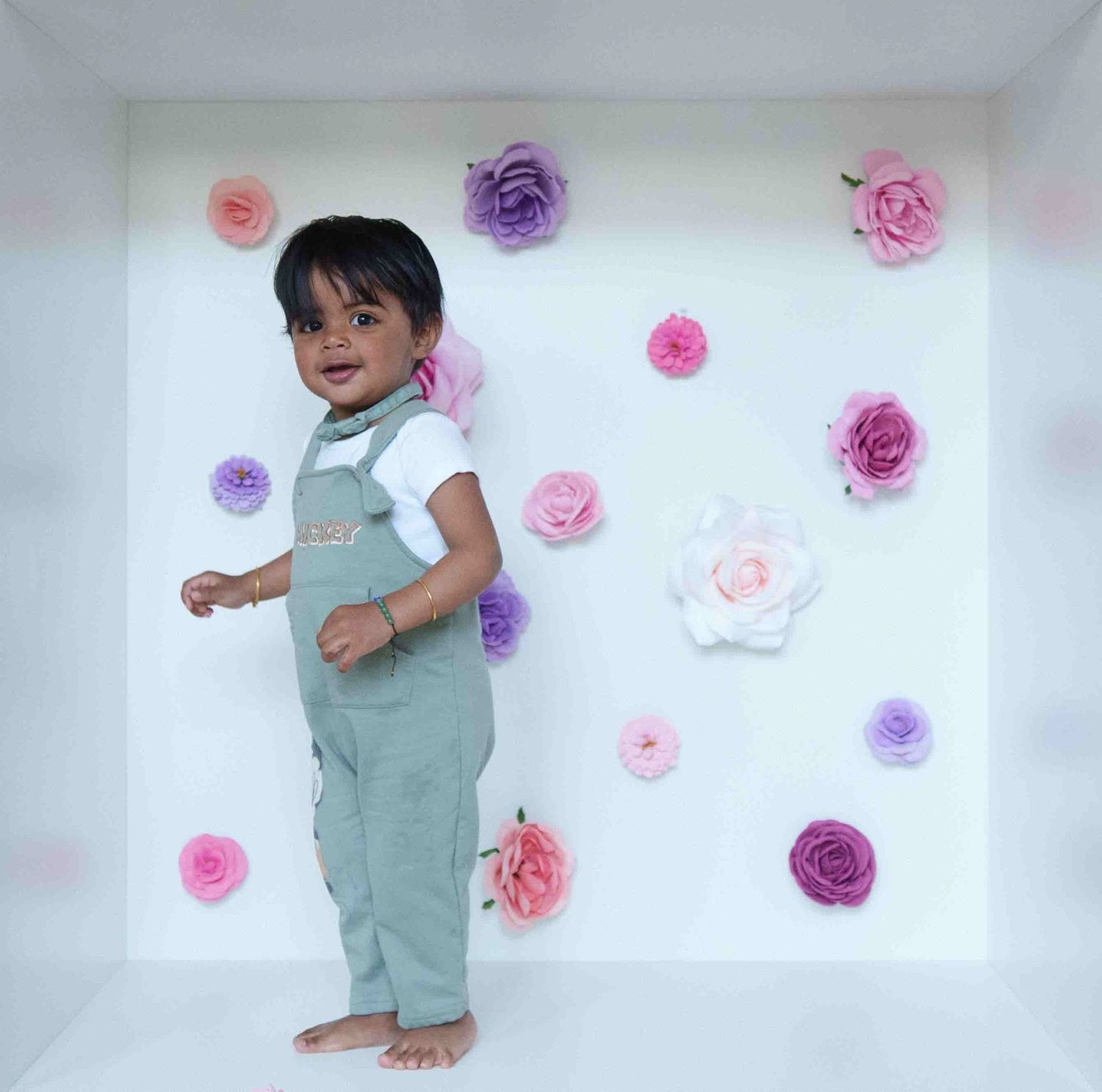 Baby wearing mint green overalls poses against white wall decorated with pink and purple paper flowers.