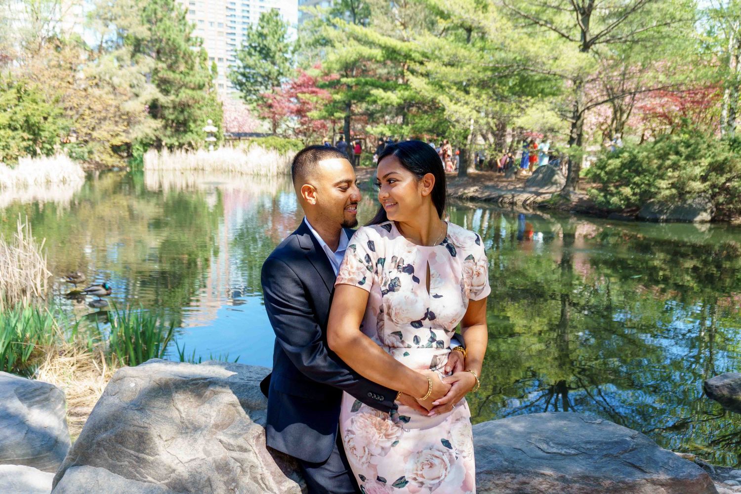 A couple embraces by a tranquil pond surrounded by spring foliage.