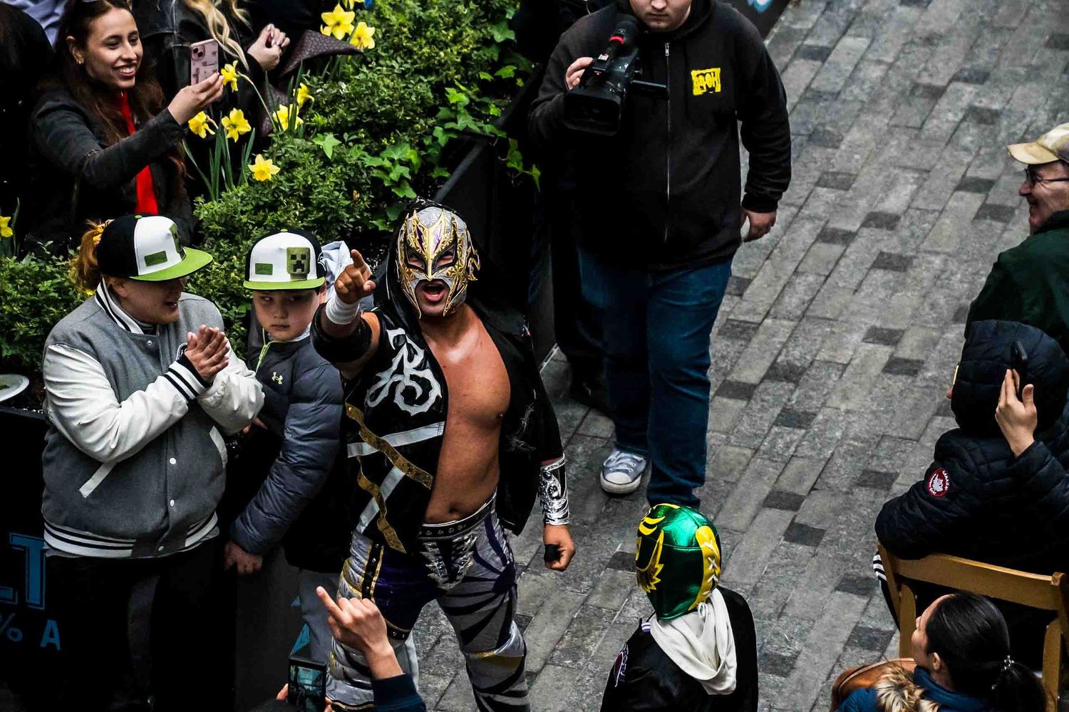 Masked wrestler greeting fans at an outdoor wrestling event with spectators watching and taking photos.