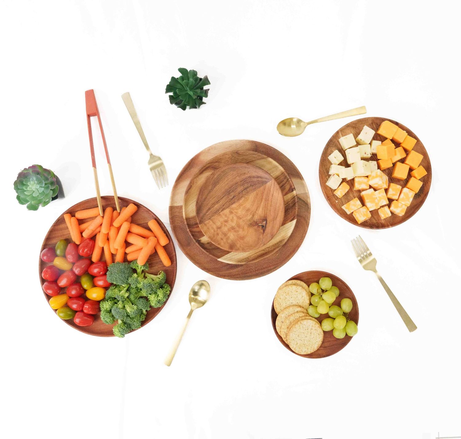 Acacia wood serving boards with fresh vegetables, cheese, crackers, and grapes, styled with gold and red utensils on white.
