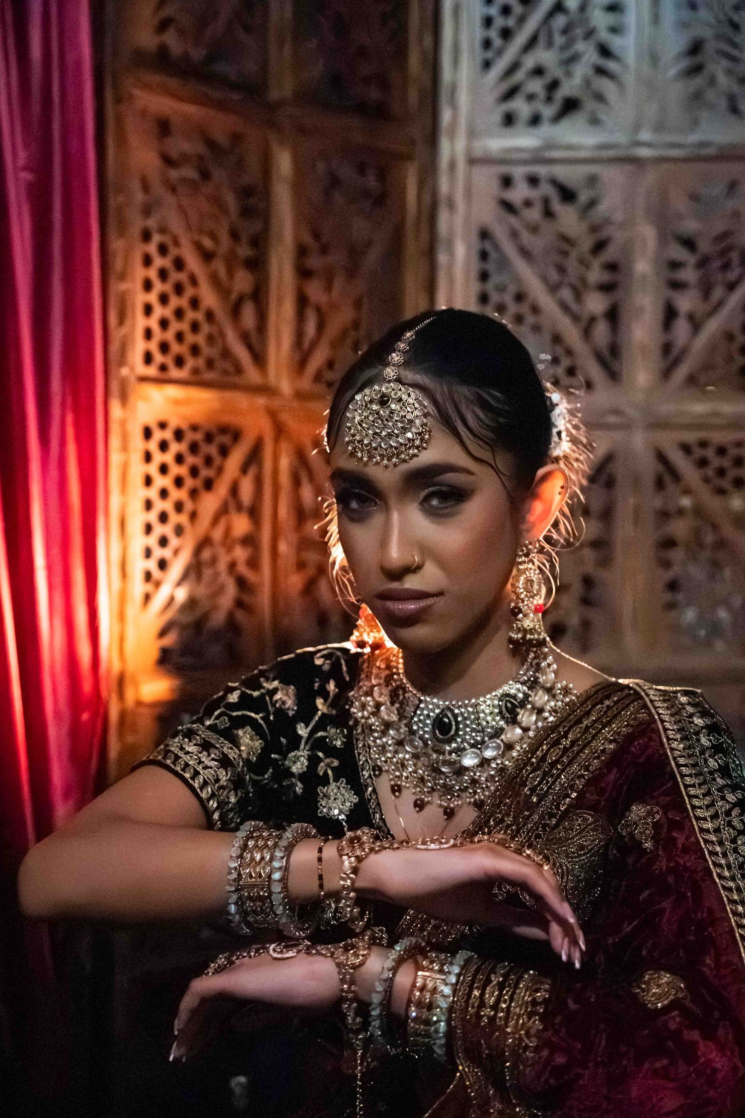 Traditional Indian bridal attire with ornate jewelry and embellishments against an intricately carved backdrop with warm lighting.