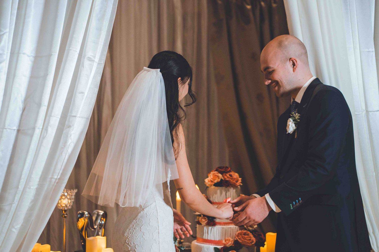 A bride and groom participate in a unity candle ceremony during their wedding with white drapes in the background.