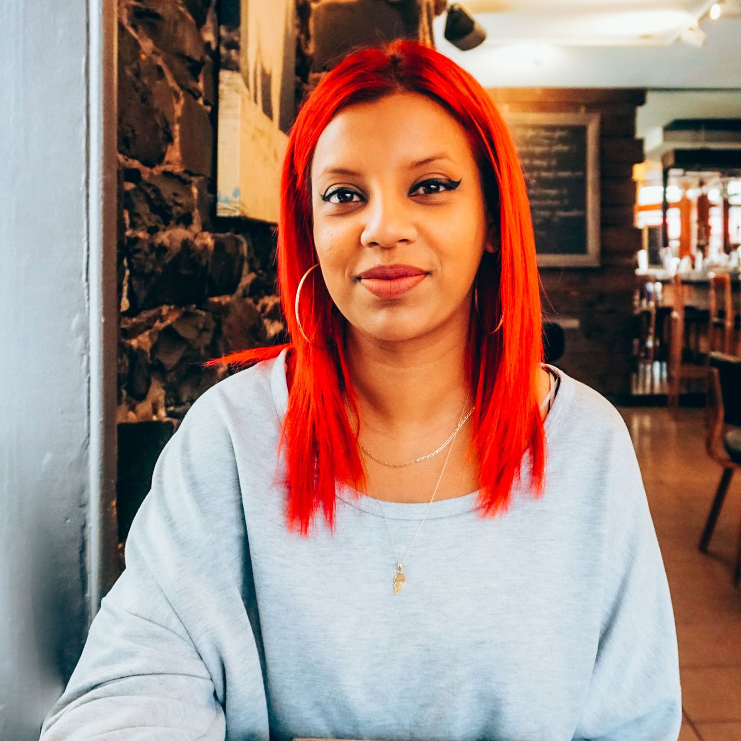 Woman in grey sweatshirt with bright red hair poses in casual indoor setting.