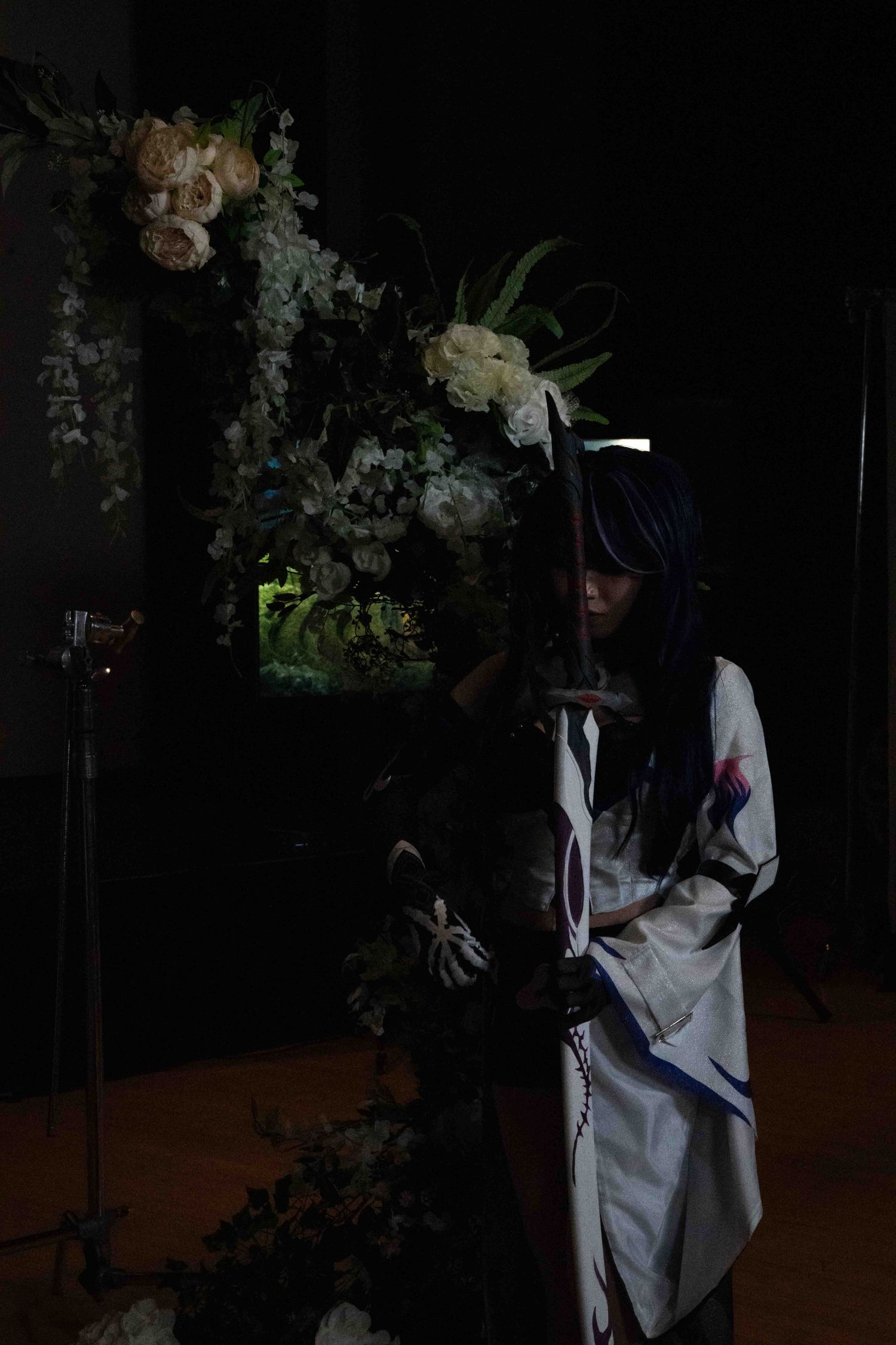 Dark atmospheric scene with figure in white clothing among floral arrangements.