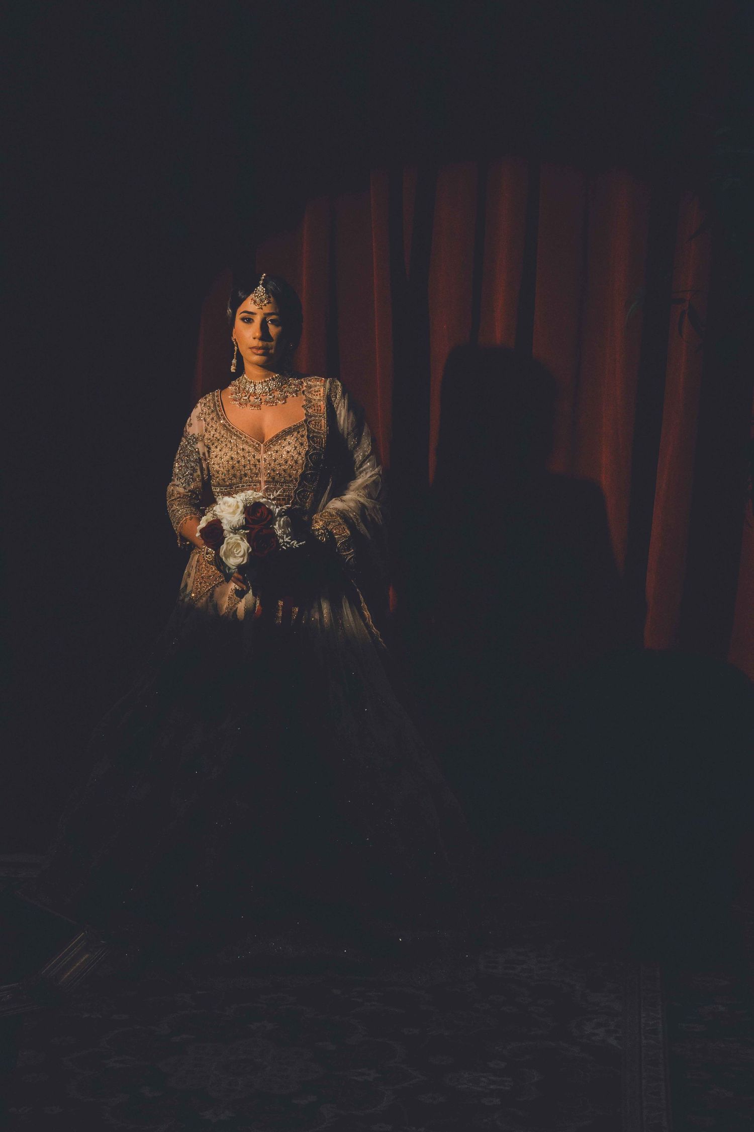 Moody portrait of a bride in an ornate dress against dark red curtains with dramatic lighting.