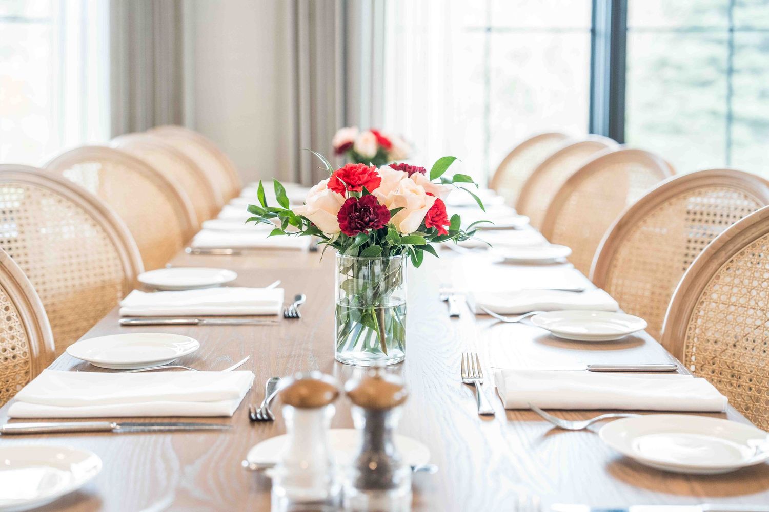 Elegant long dining table set for a formal dinner with wicker chairs and a floral centerpiece of red and white roses.