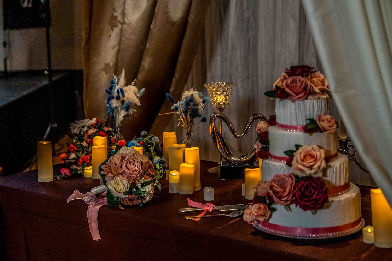 Moody still life composition of pink roses and candles arranged on wooden table with dramatic lighting and shadows.