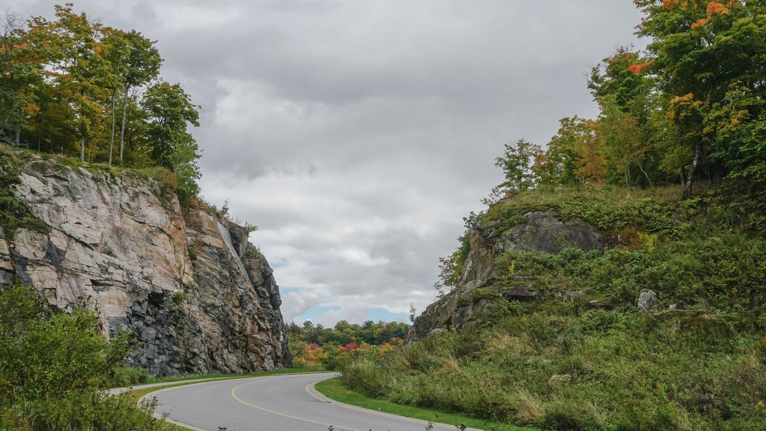 Winding mountain road through autumn trees with dramatic cloudy skies overhead.