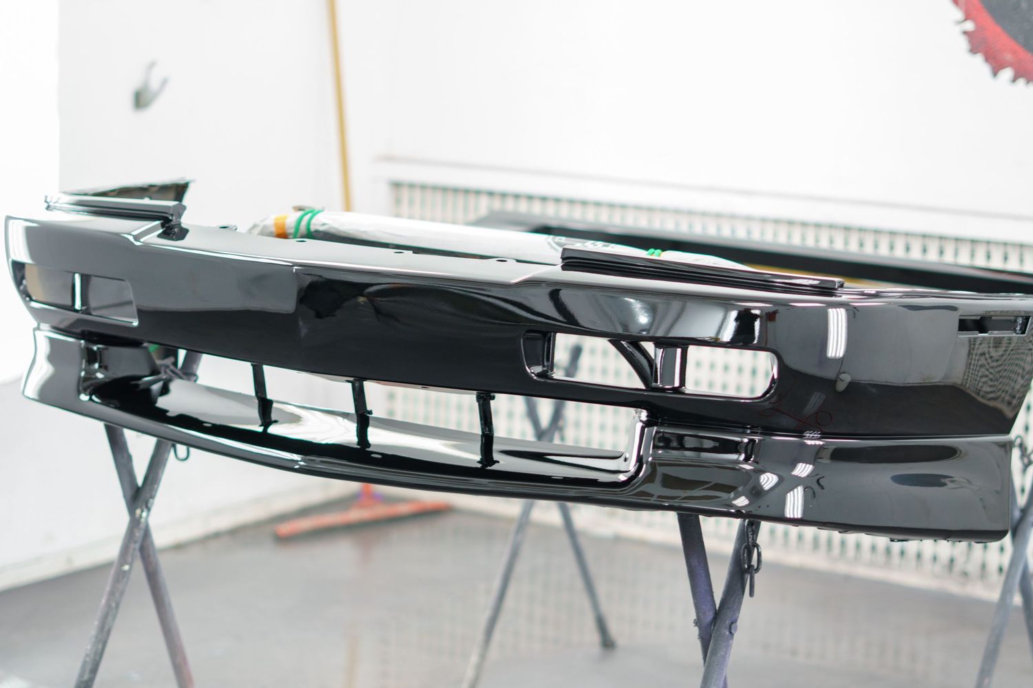 Shiny black automotive front bumper with grille on a workshop stand in a paint booth.