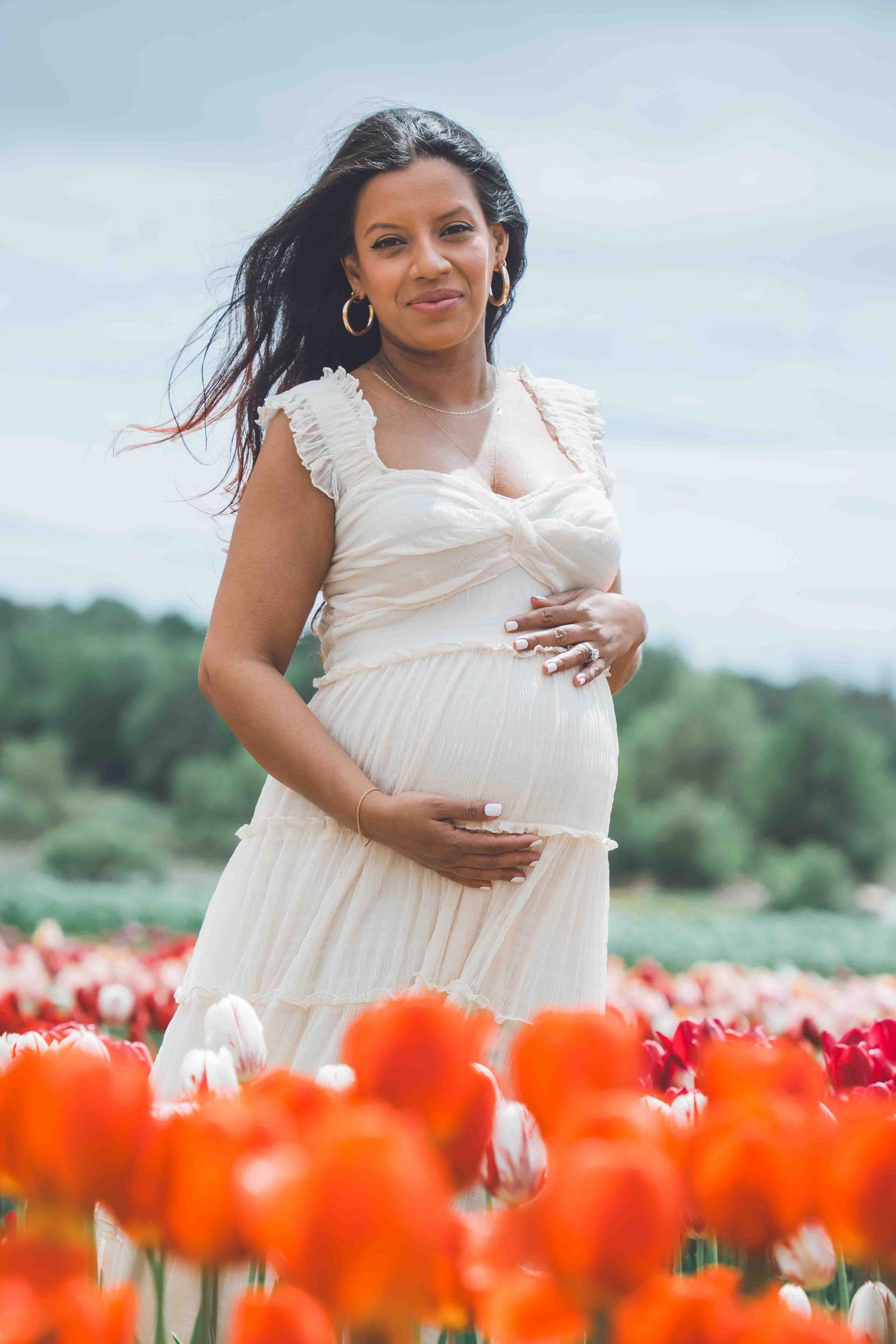 Expectant mother in white dress posing among vibrant orange tulips.