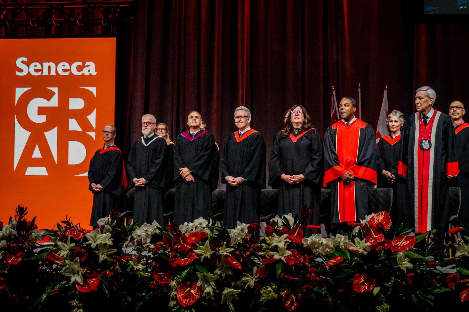 Graduates in black robes and caps seated during commencement ceremony with Seneca branding visible.