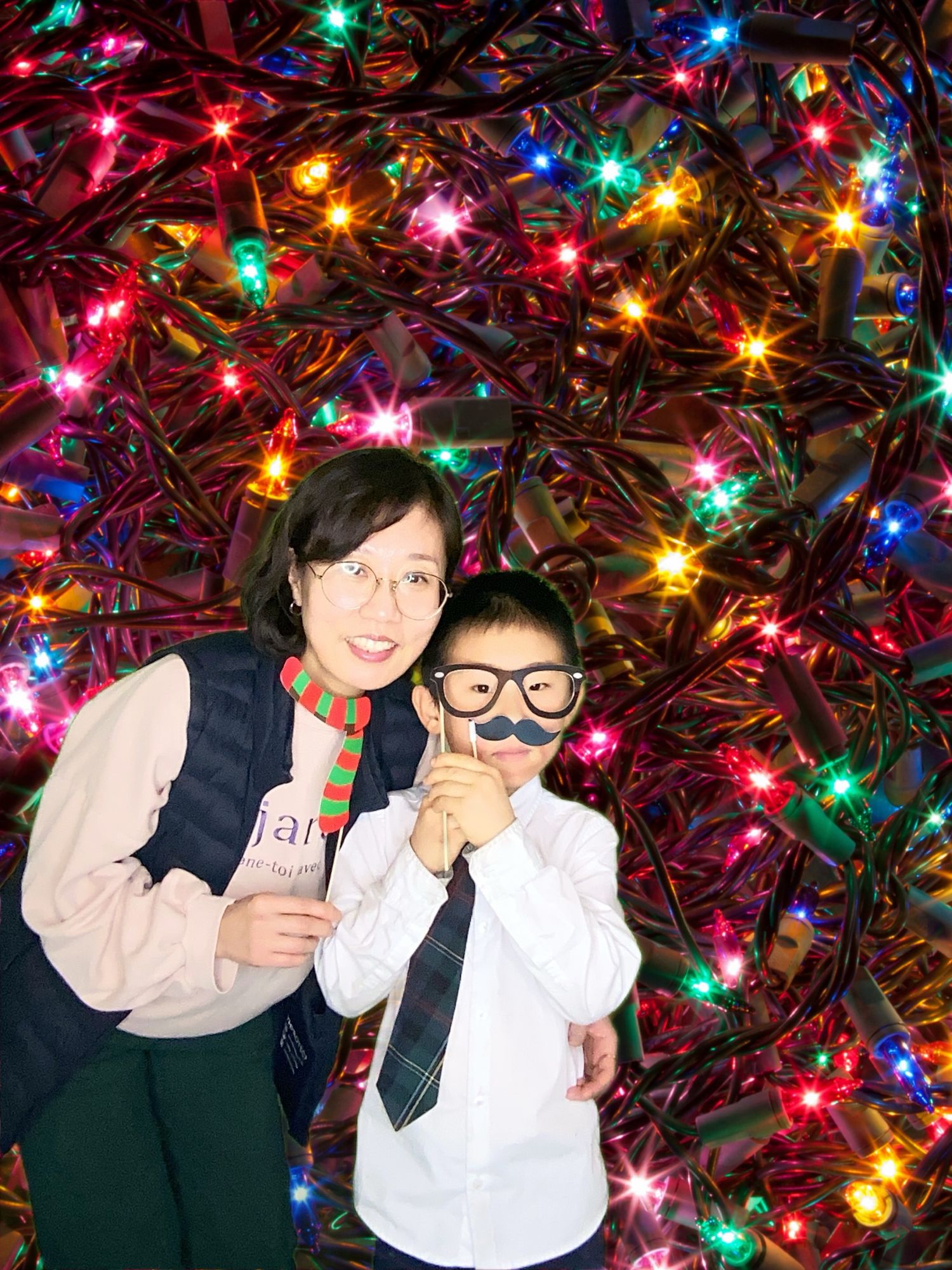 Two people pose with props against a colorful Christmas light background.