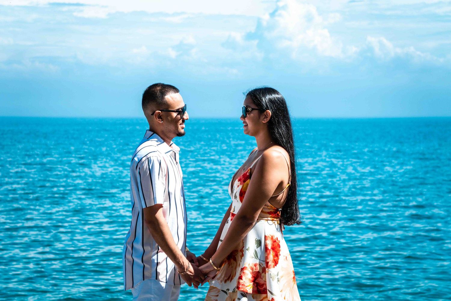 A couple stands together against a vibrant turquoise ocean backdrop on a sunny day.