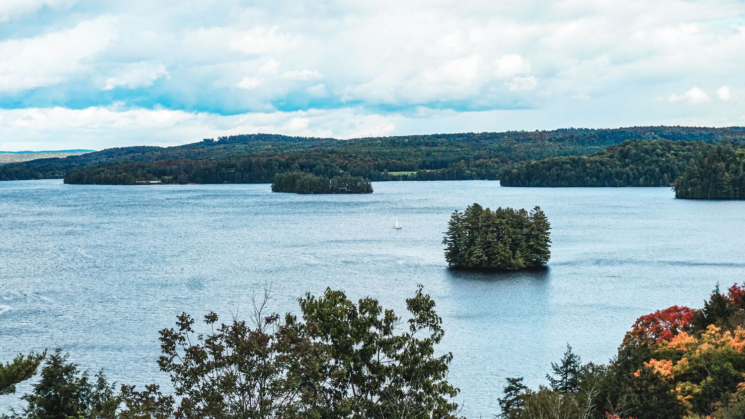 Panoramic view of a lake surrounded by forested hills and islands under cloudy skies.