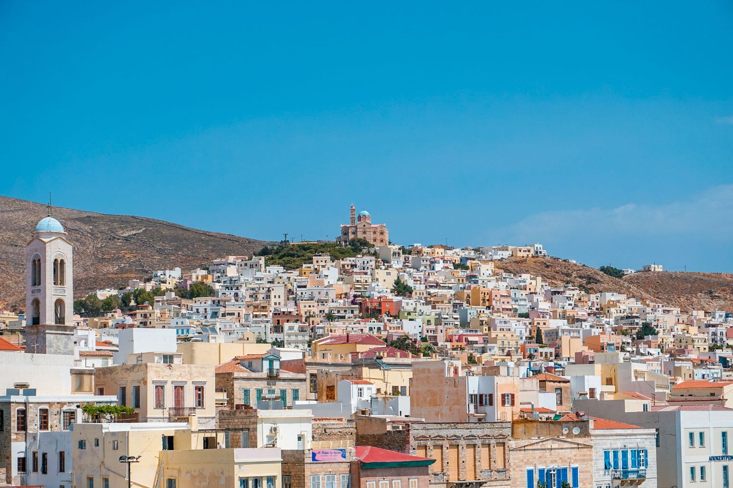 Hillside Greek town with white buildings and church dome against bright blue sky.