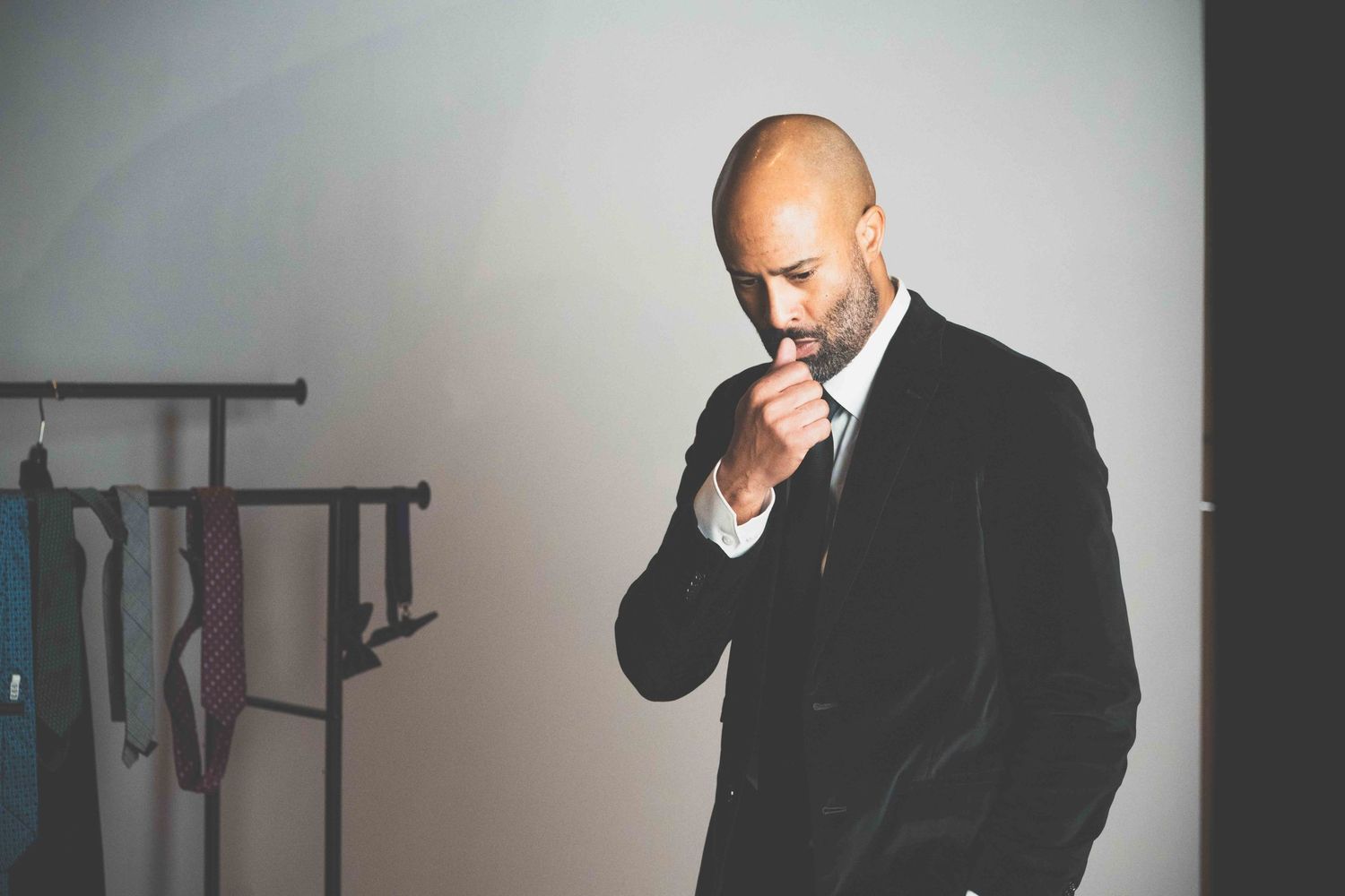 A person in a black suit adjusts their tie while getting ready in a dimly lit room.