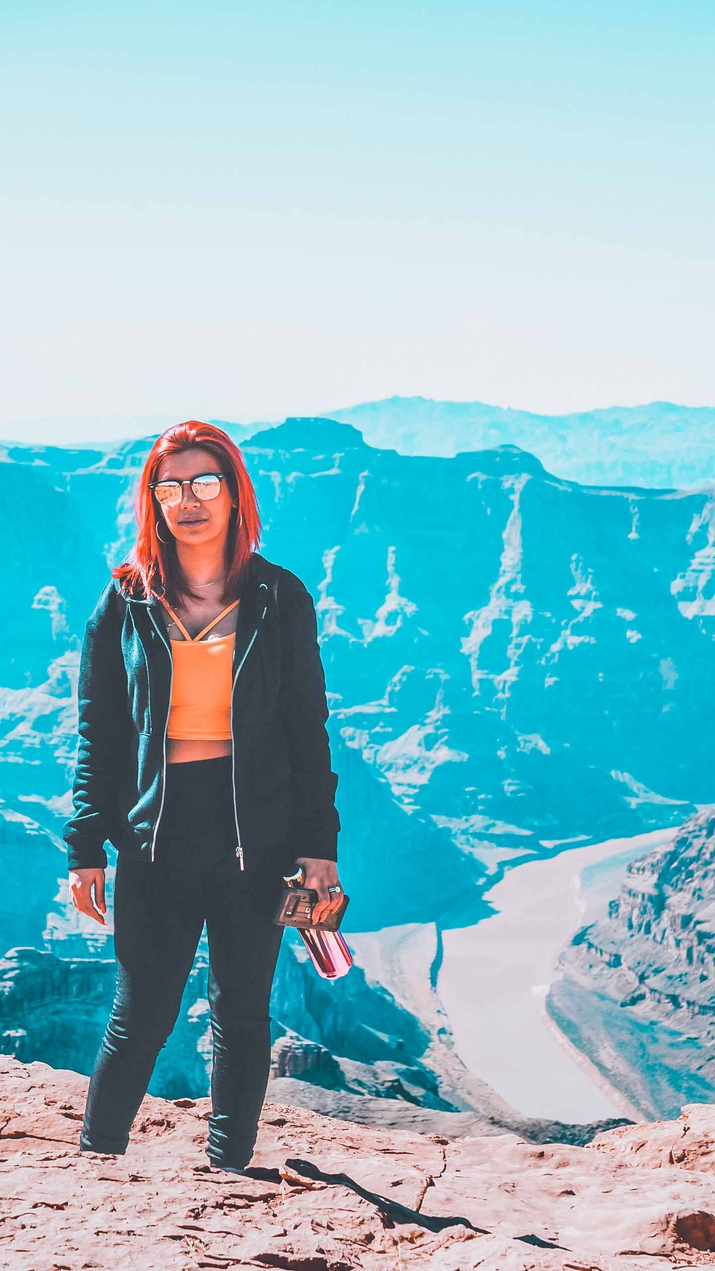 Person in black jacket and sunglasses standing on mountain overlook with dramatic blue canyon view.