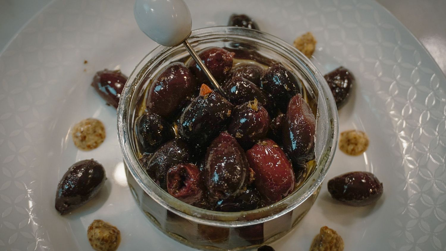 Glass jar containing dark marinated olives and antipasto on a white plate.