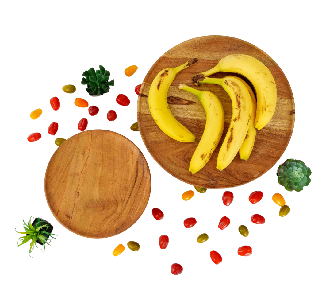 Overhead view of ripe bananas on a wooden board surrounded by colorful dried fruits and small succulents on white background.