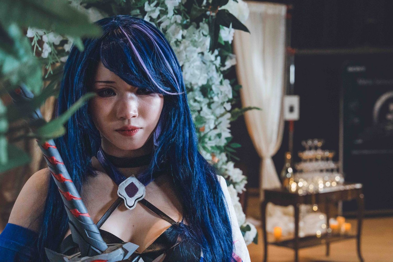 Person with vibrant blue hair poses among indoor plants and white curtains.