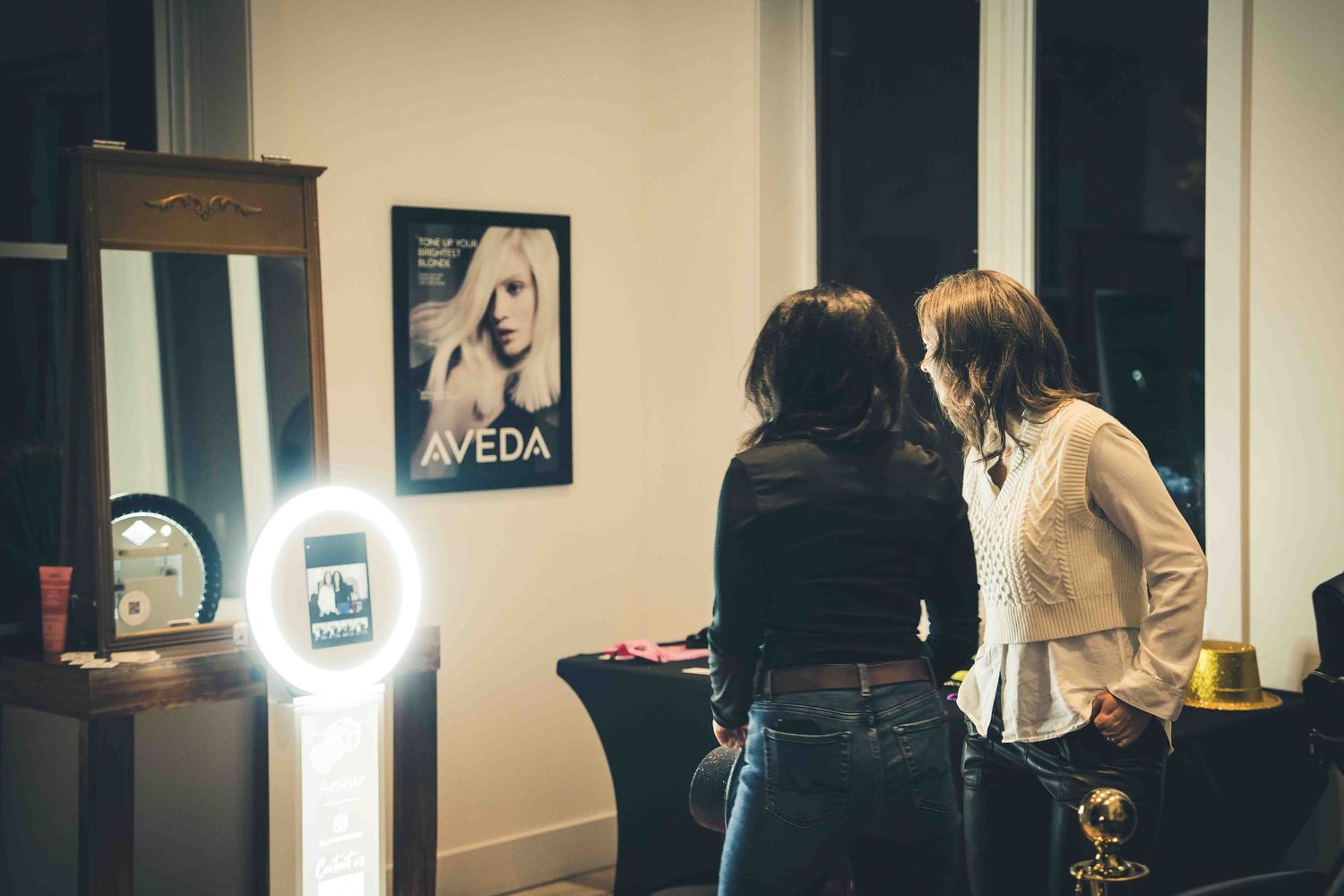Event guests viewing photo booth display with illuminated screen and Aveda poster.