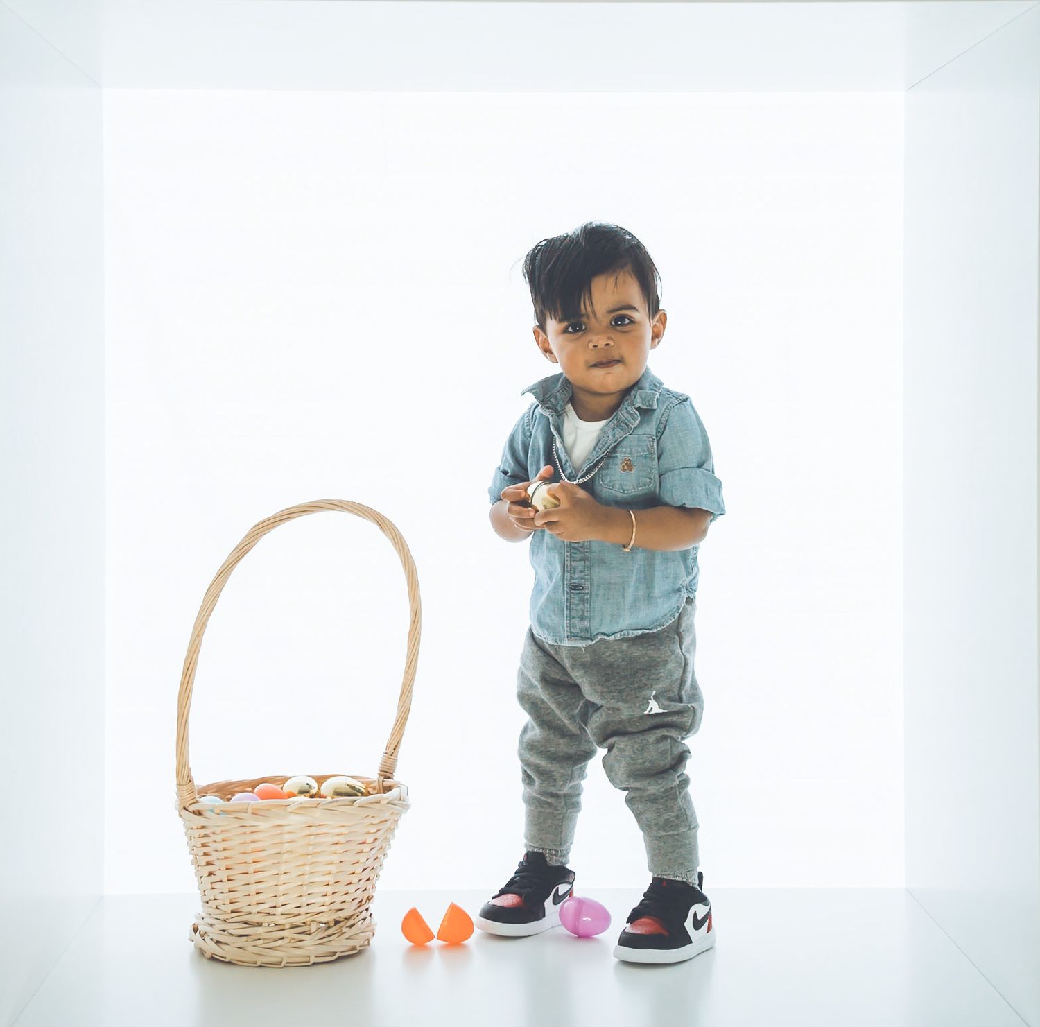 Child in casual outfit stands next to wicker Easter basket on white background.