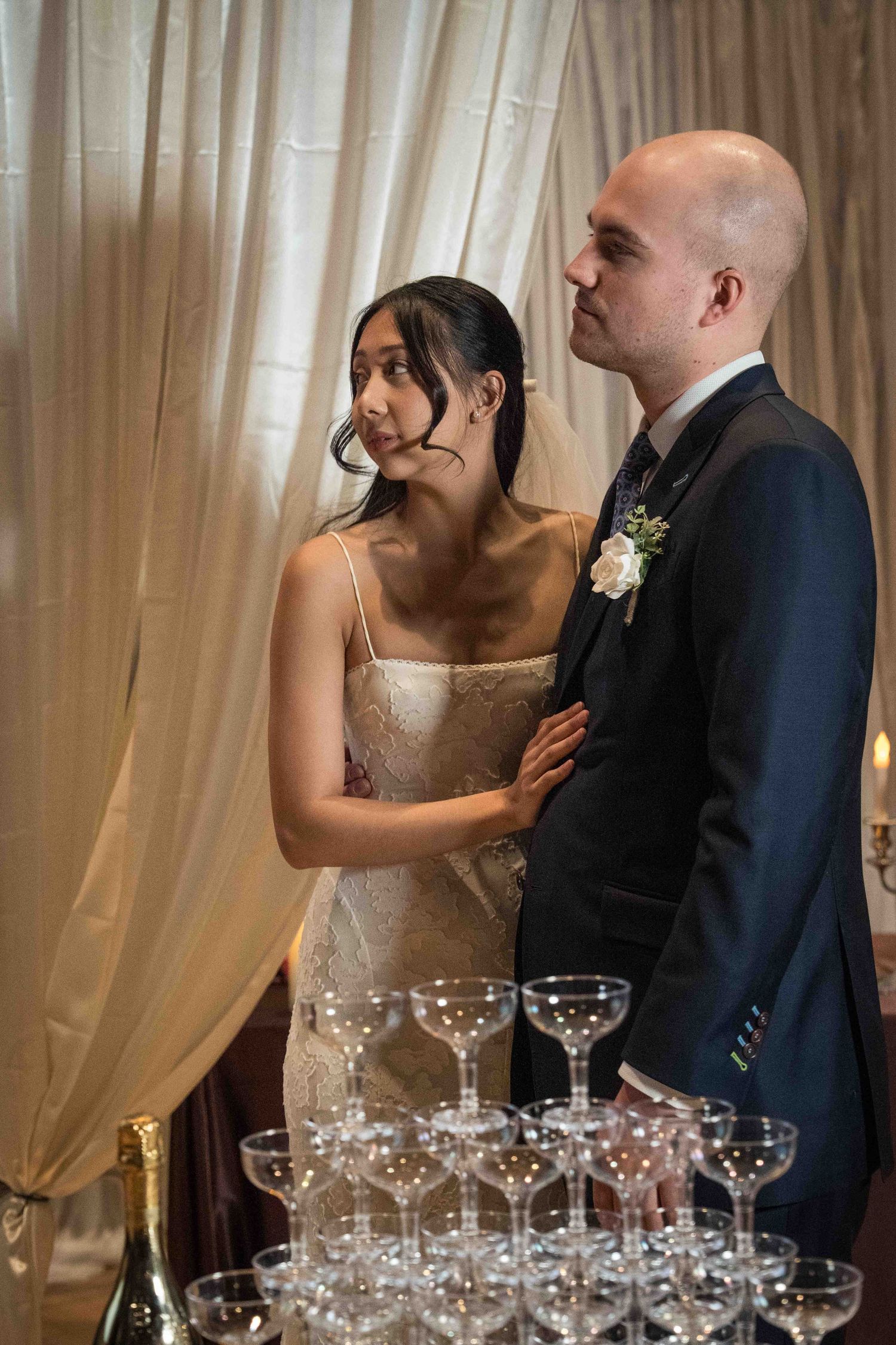 Elegant couple stands beside a tower of champagne glasses at their wedding reception.