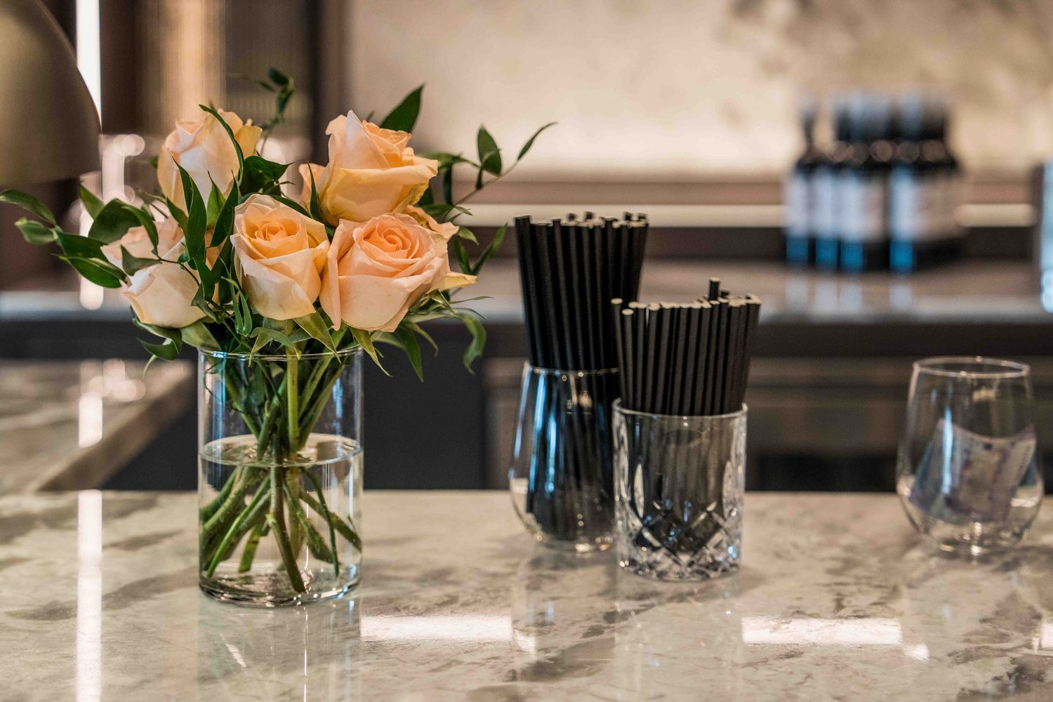 Elegant marble countertop with peach roses in glass vase, black straws in crystal holder, and glassware in modern kitchen.