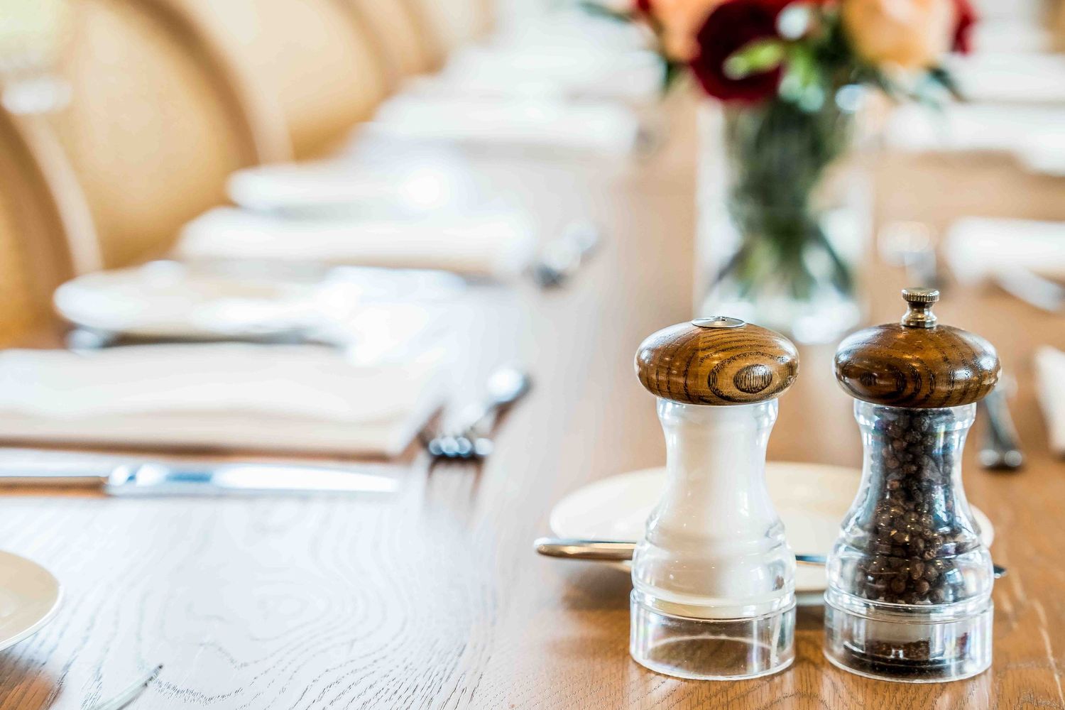 Salt and pepper mills on a elegantly set restaurant dining table with cutlery and floral centerpiece.