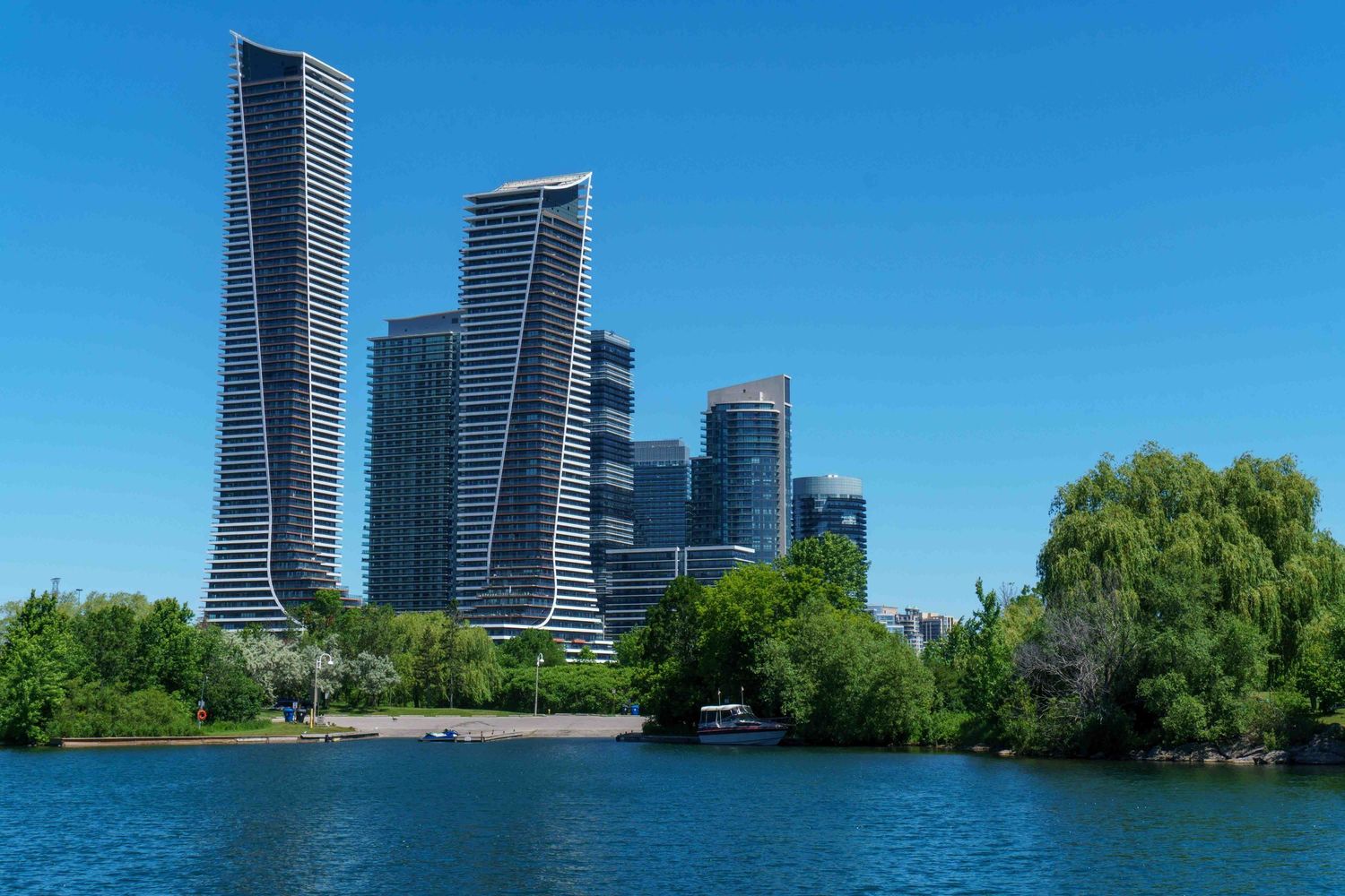Modern glass skyscrapers along Toronto's waterfront with Lake Ontario and trees in view.