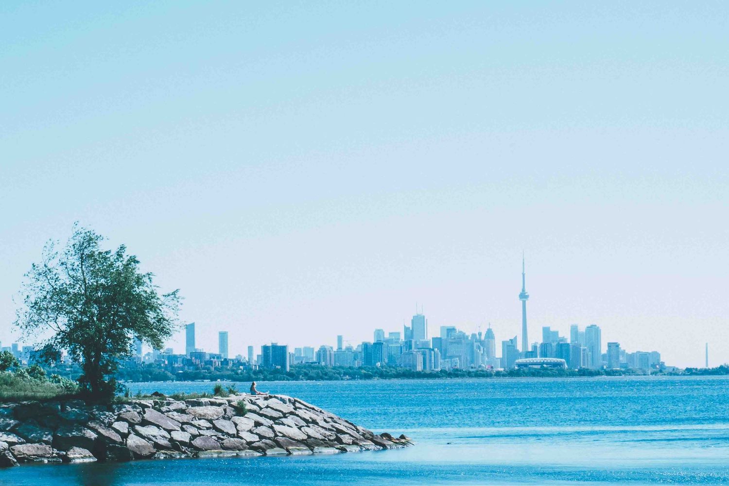 Toronto's urban skyline viewed across Lake Ontario with rocky shoreline in foreground.