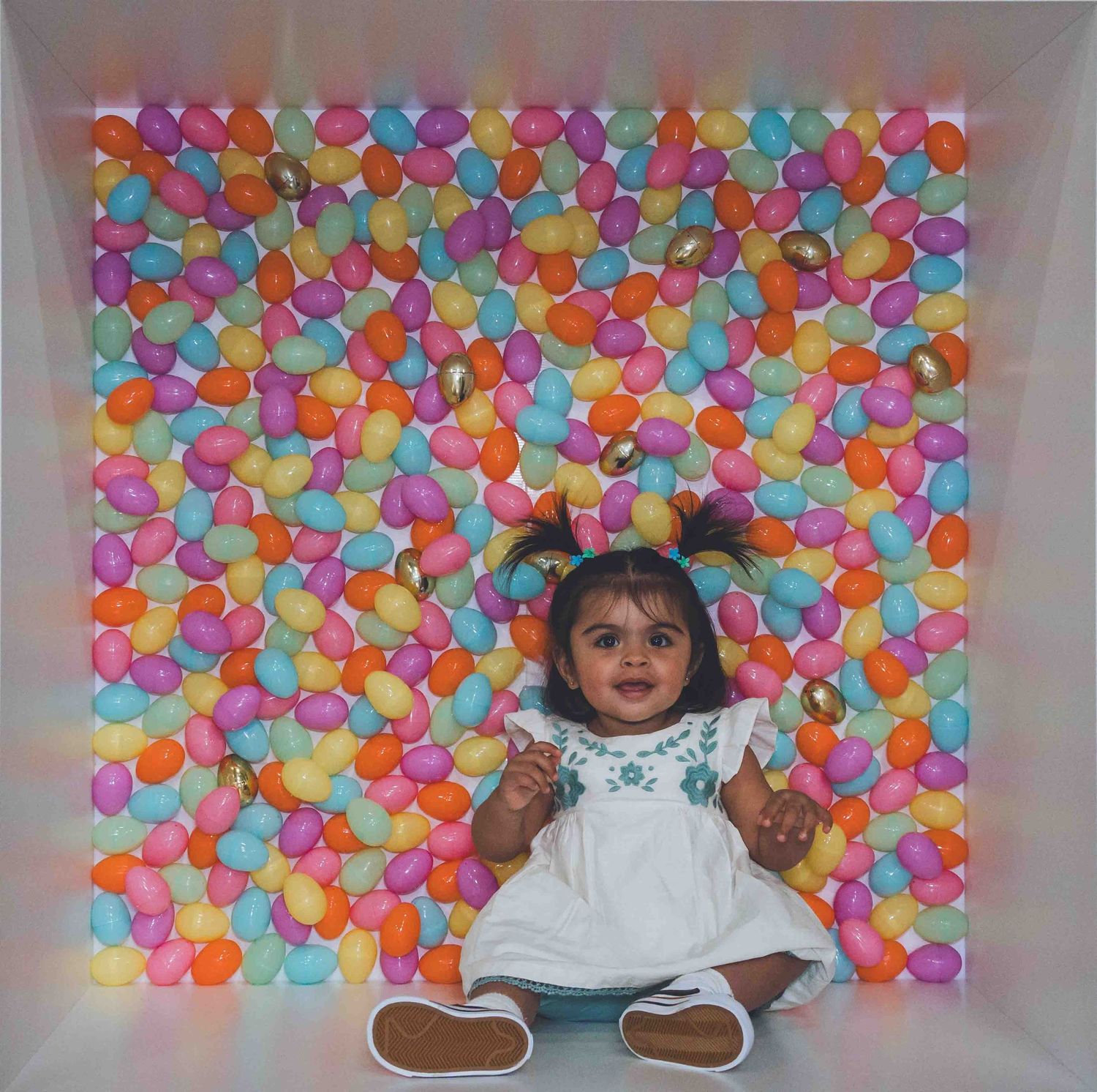 Baby sits in front of vibrant rainbow balloon wall wearing white dress with floral details.