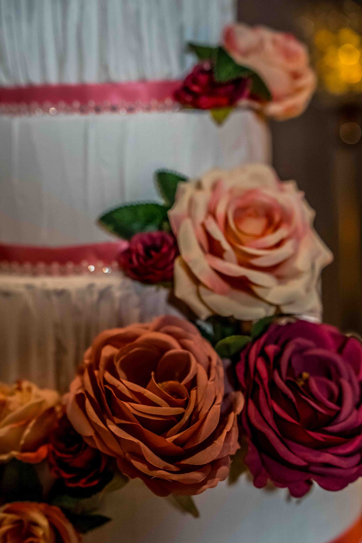 Close-up of elegant roses in burgundy, peach and pink colors decorating a white wedding cake with soft bokeh lighting.