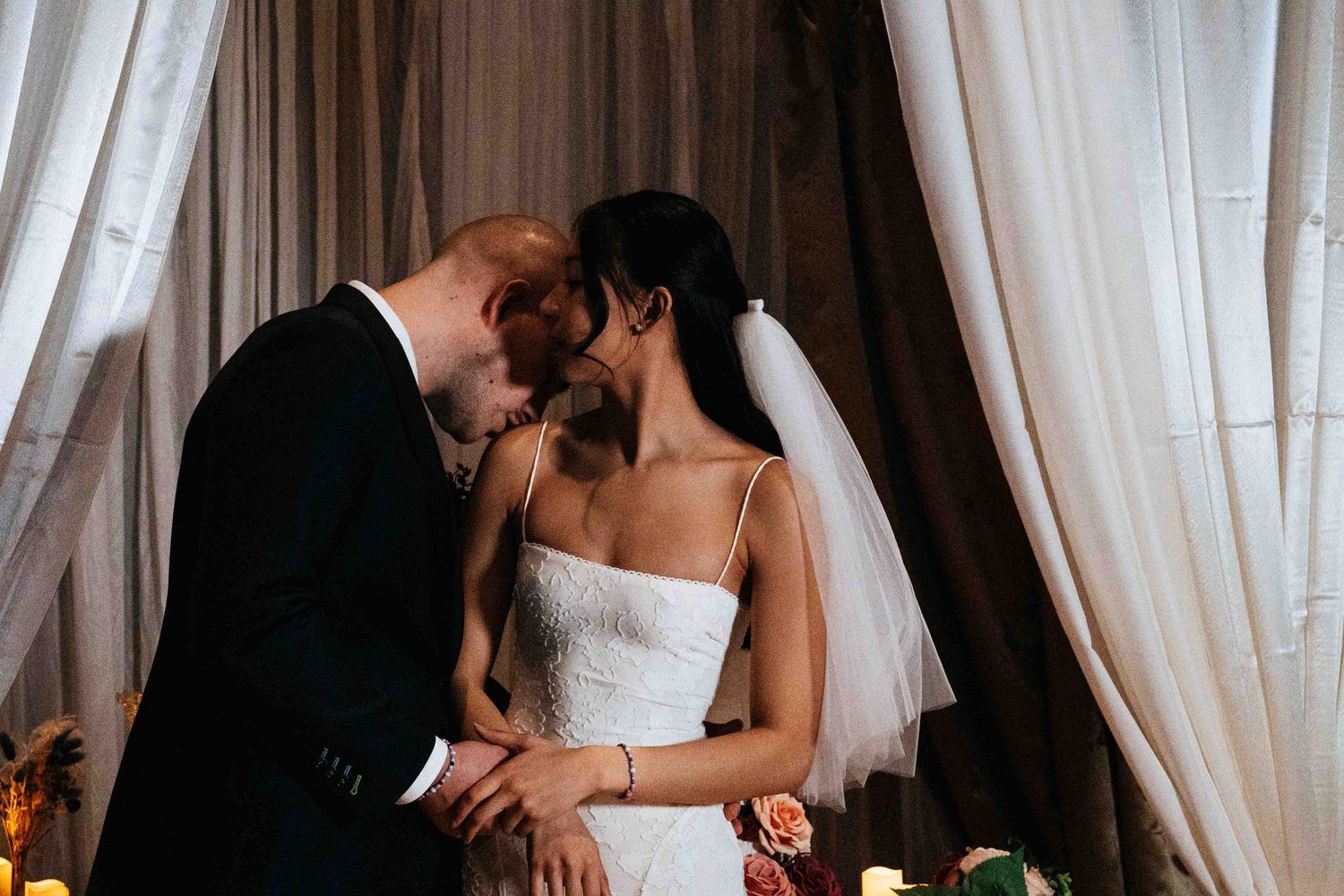 A romantic moment between newlyweds sharing a kiss near white curtains during their wedding reception.