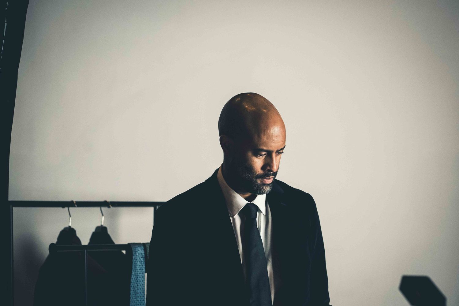 A moody portrait in dark lighting shows a figure in a black suit and tie standing against a light wall.