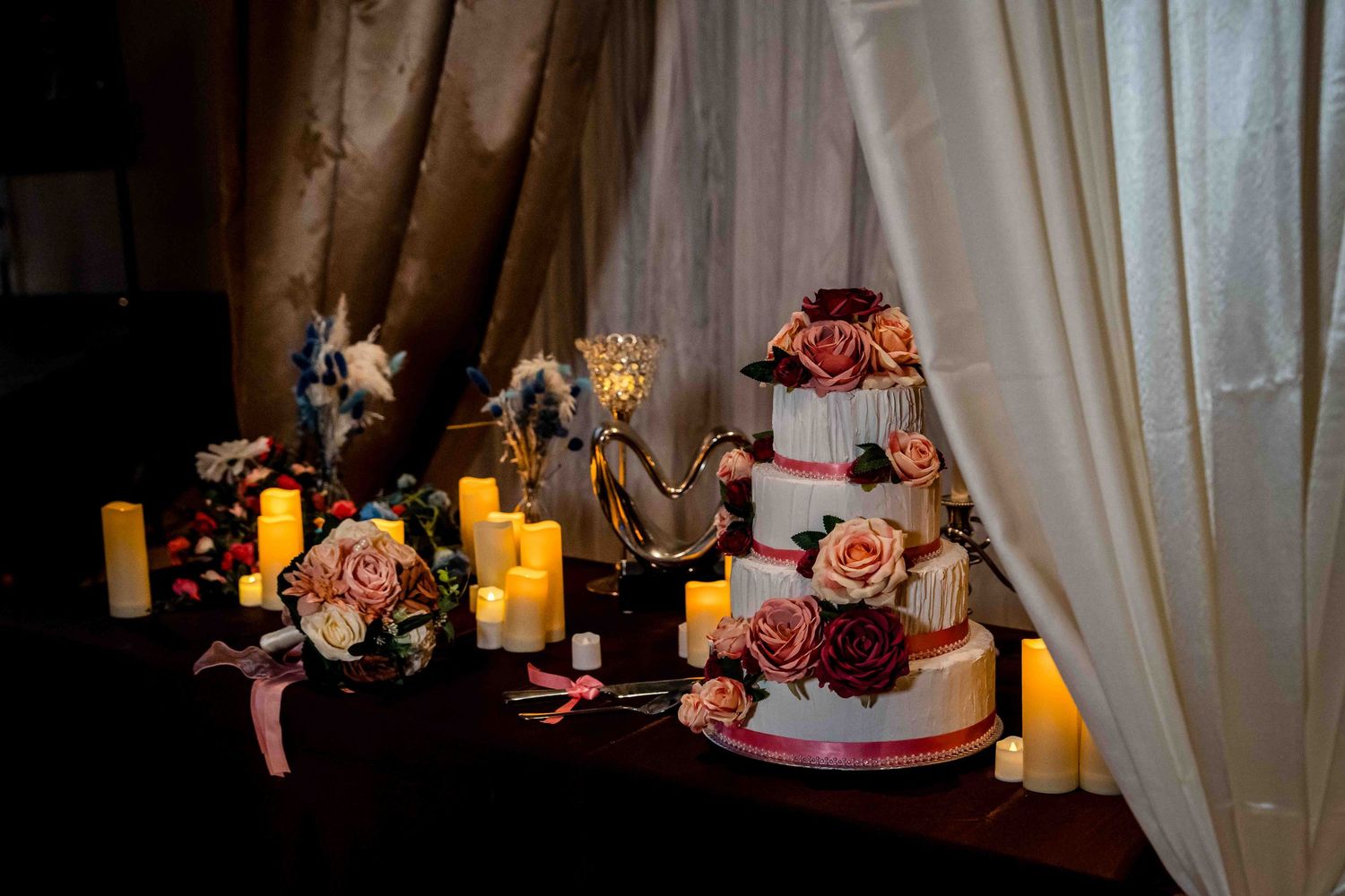 A romantic table setting with lit candles and pink roses creates an elegant atmosphere against sheer curtains.